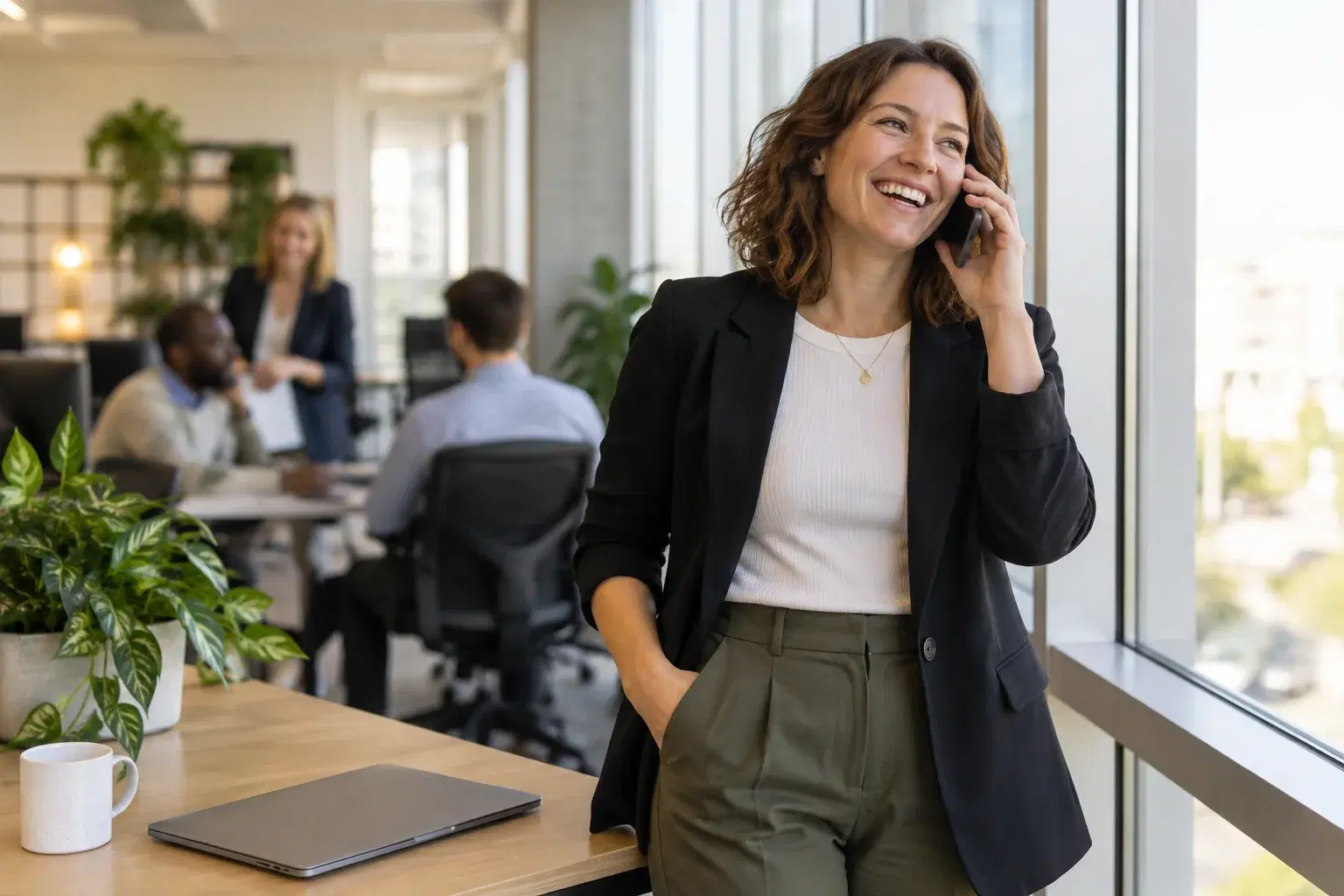 A staffing agency recruiter standing by an office window, laughing on a phone call with a candidate, laptop closed on the desk beside her